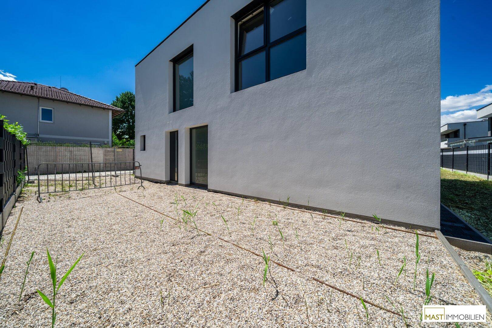Moderne weiße Fassade eines Neubaus mit großen Fenstern und einem Kiesgarten unter blauem Himmel.
