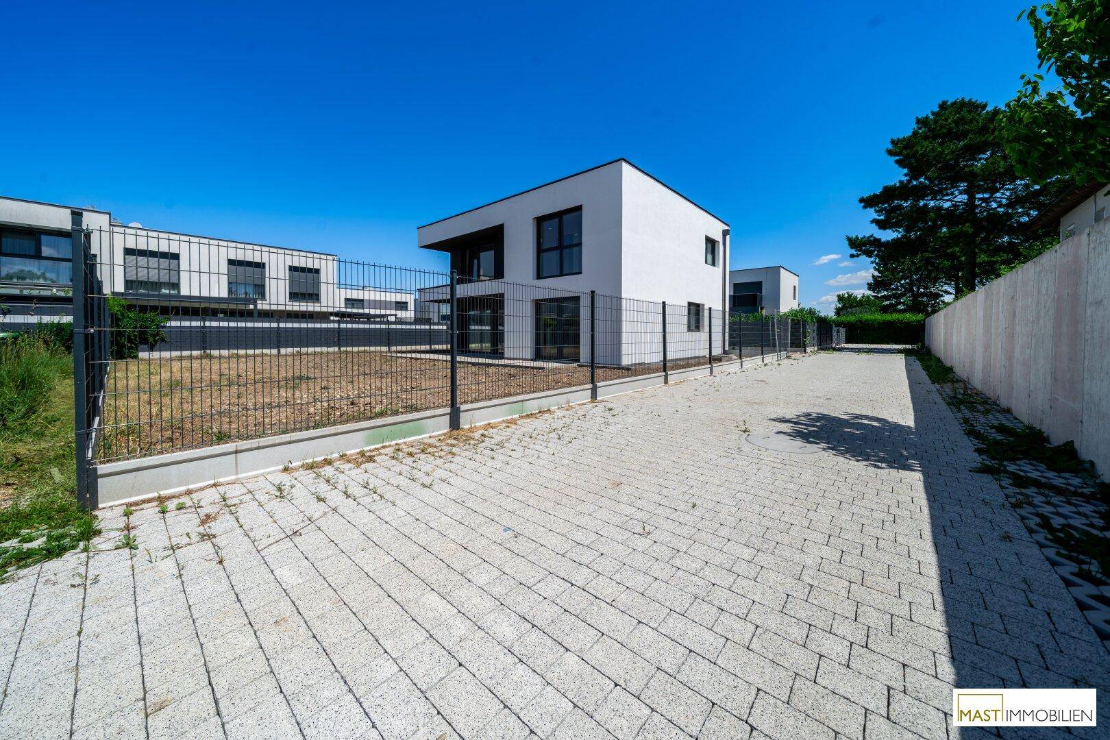 Gepflasterte Zufahrt zu einem modernen Neubau mit umlaufendem Zaun und angrenzendem Grünbereich.
