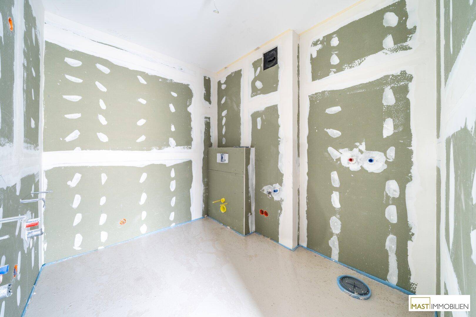 Unfertiges Badezimmer im Rohbau mit sichtbaren Sanitärinstallationen und feuchtigkeitsbeständigen Wänden.