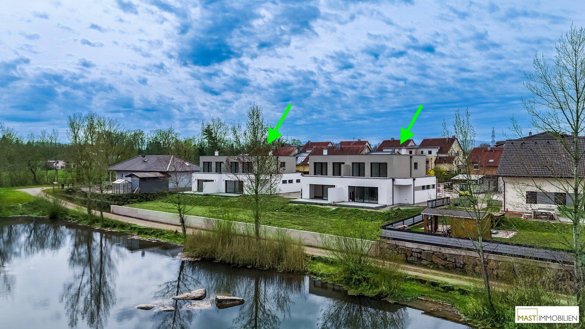 Zwei moderne Häuser am Ufer eines Teiches, umgeben von grüner Landschaft unter bewölktem Himmel.
