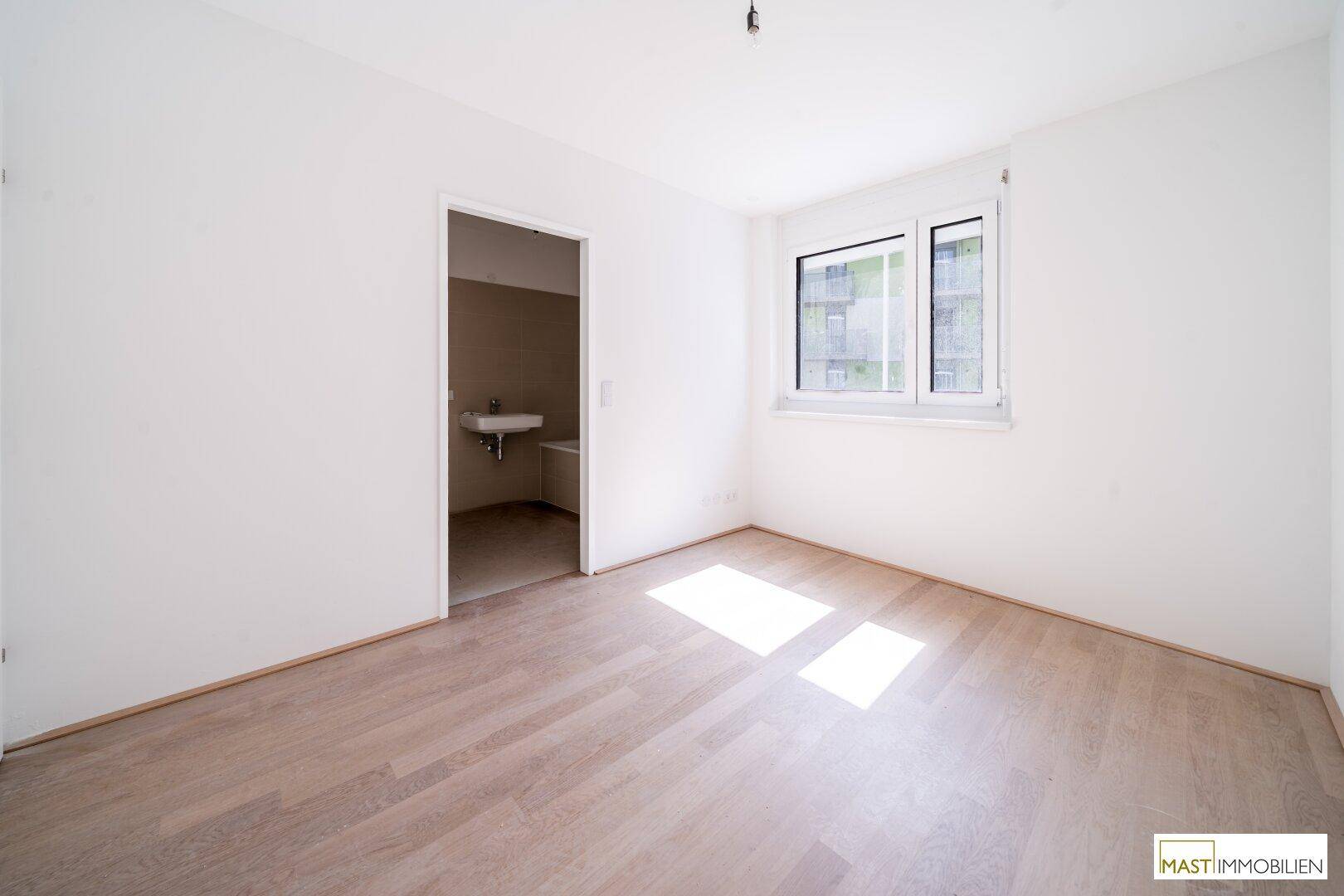 Schlafzimmer mit hellem Holzboden, großem Fenster und direktem Zugang zu einem Badezimmer.