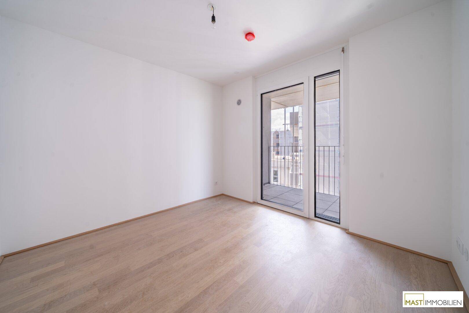 Leeres Zimmer mit Zugang zu einem Balkon und hellem Holzboden, bereit zur individuellen Einrichtung.