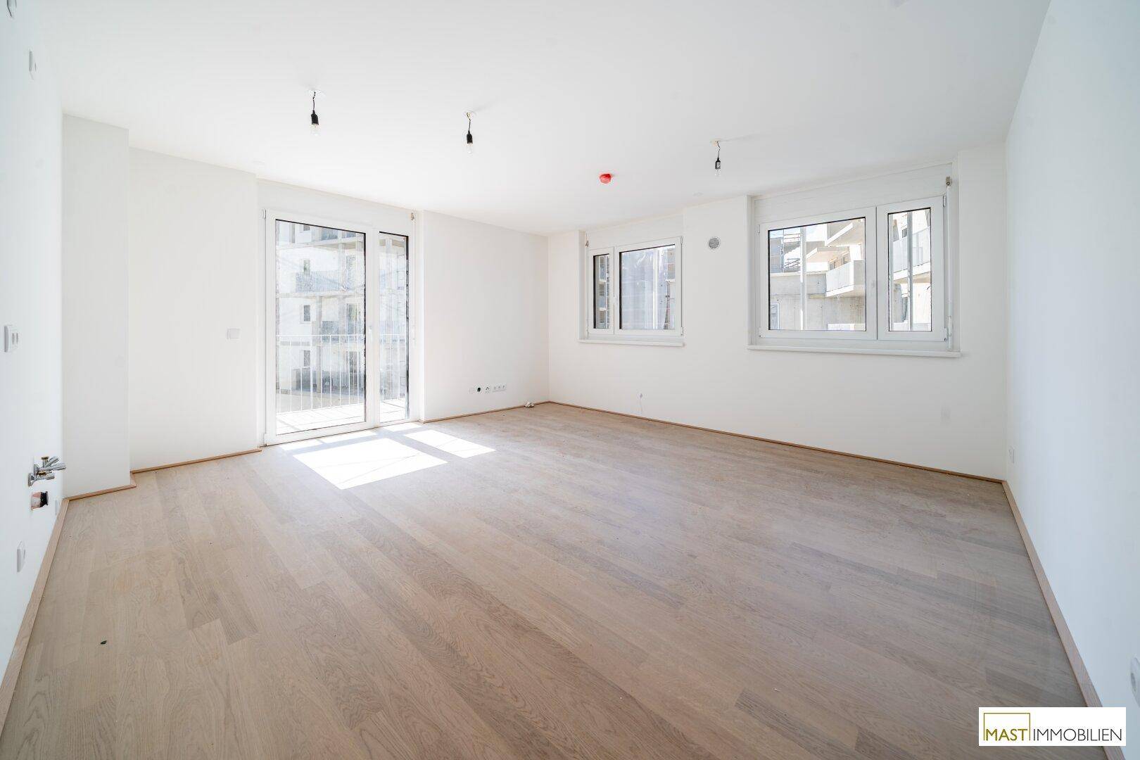 Geräumiges Wohnzimmer mit hellem Holzboden, großen Fenstern und Zugang zu einem Balkon.
