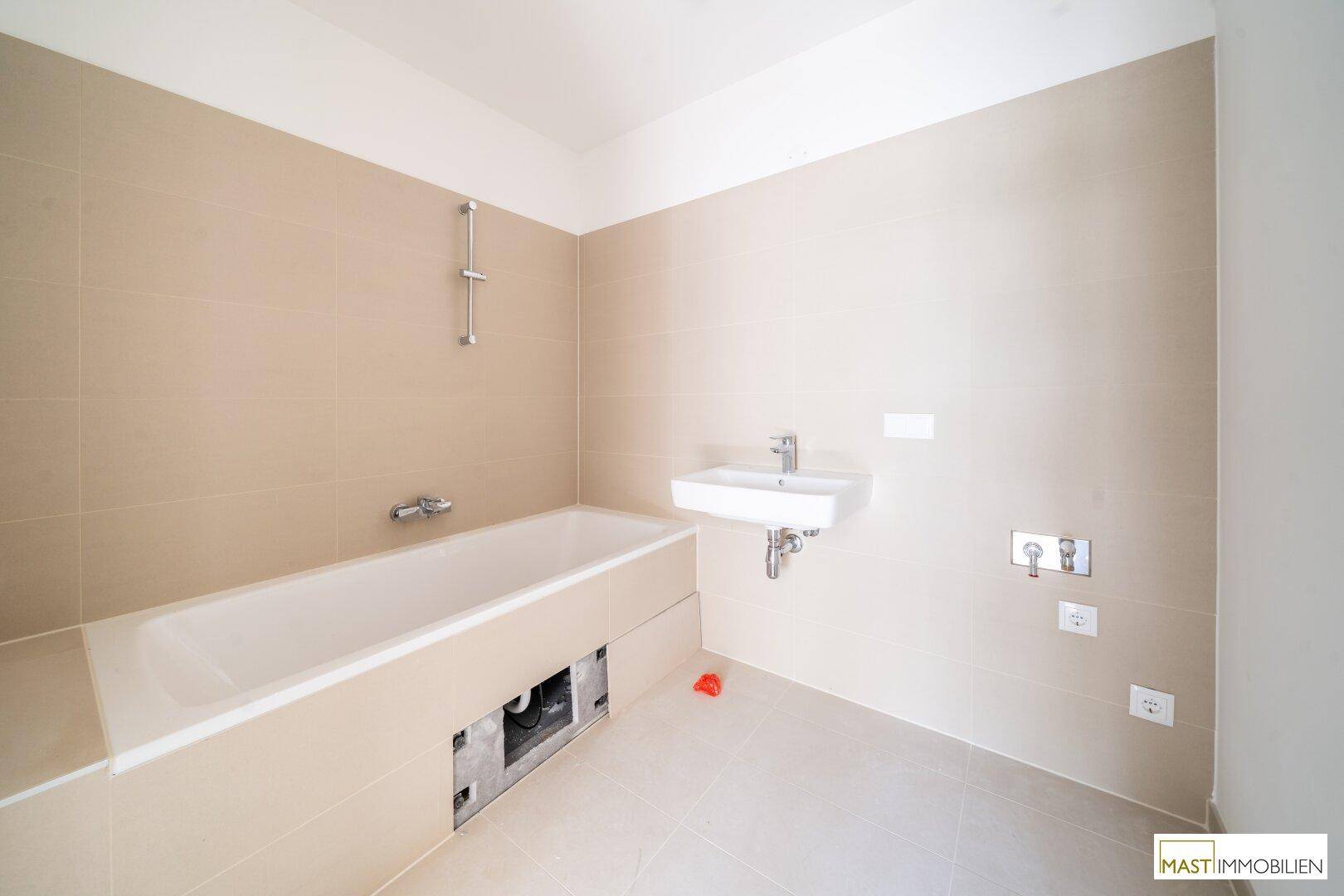 Unfertiges Badezimmer mit Badewanne, Waschbecken und beigefarbenen Fliesen an Wänden und Boden.