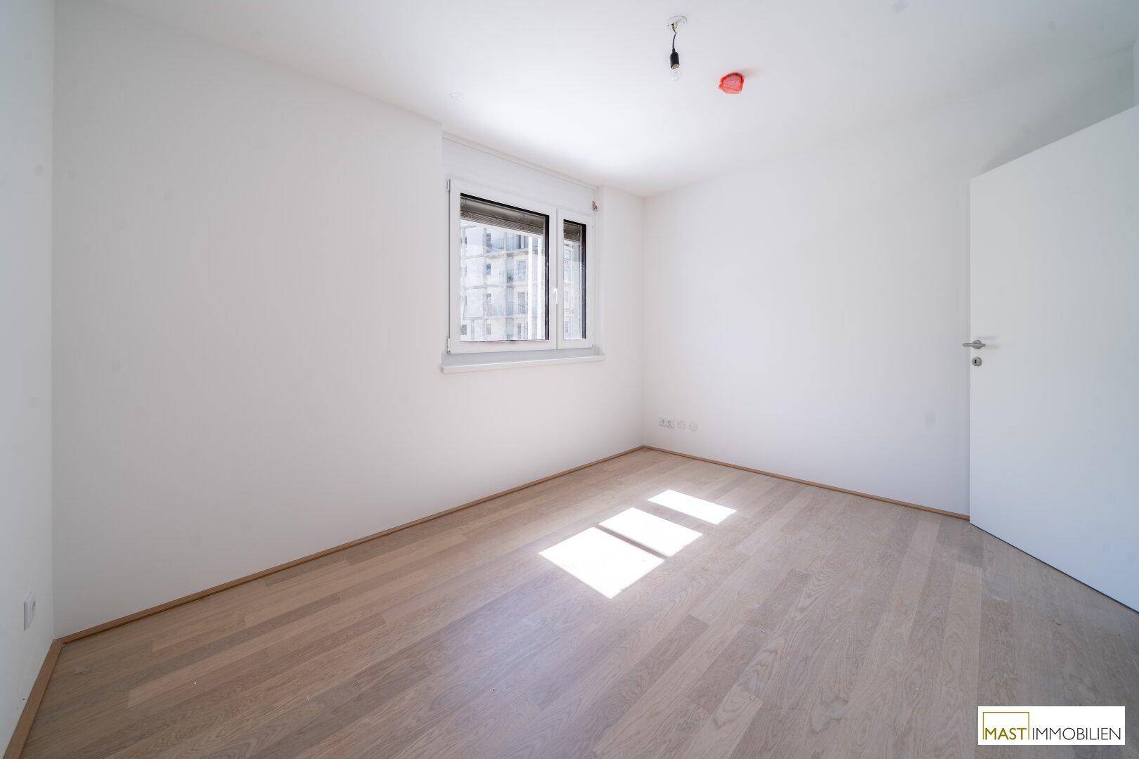 Helles Schlafzimmer mit weiß gestrichenen Wänden und hellem Holzfußboden, durchflutet von Sonnenlicht.