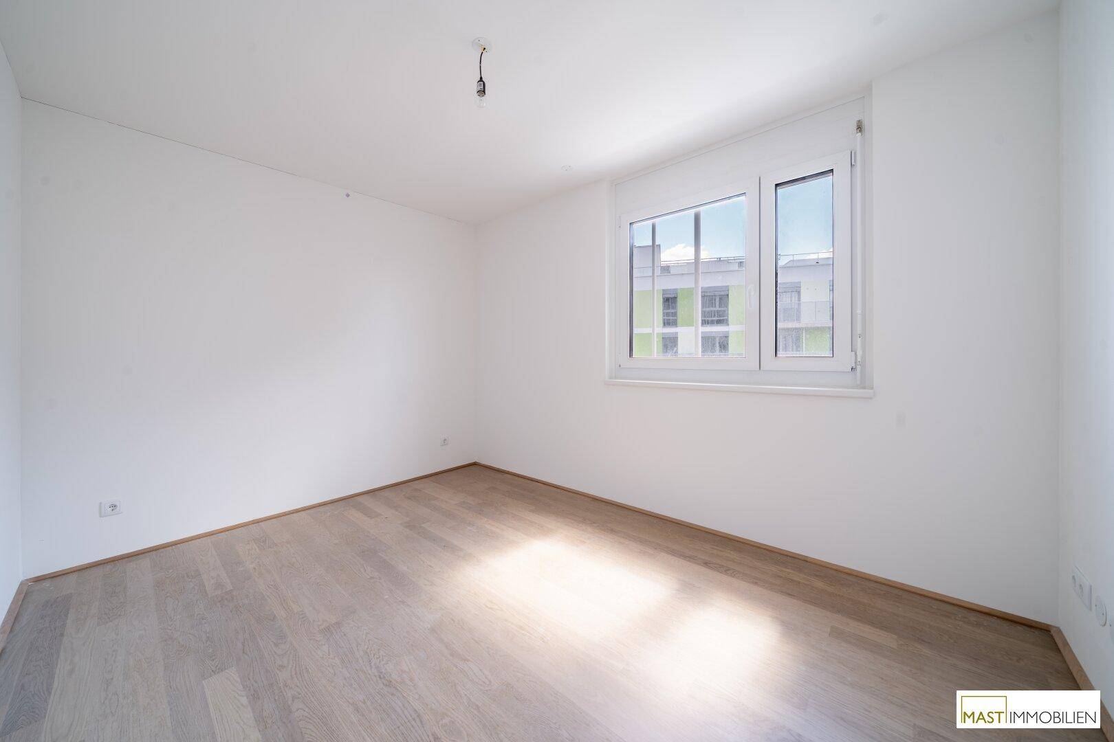 Leeres Schlafzimmer mit hellem Parkettboden und großem Fenster, das viel Tageslicht hereinlässt.