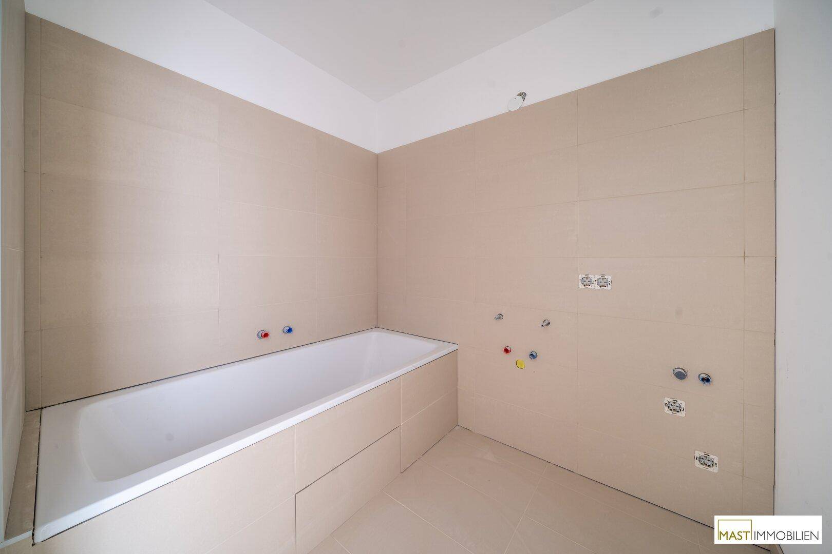 Badezimmer im Rohbau mit eingebauter Badewanne und beigefarbenen Fliesen an den Wänden.