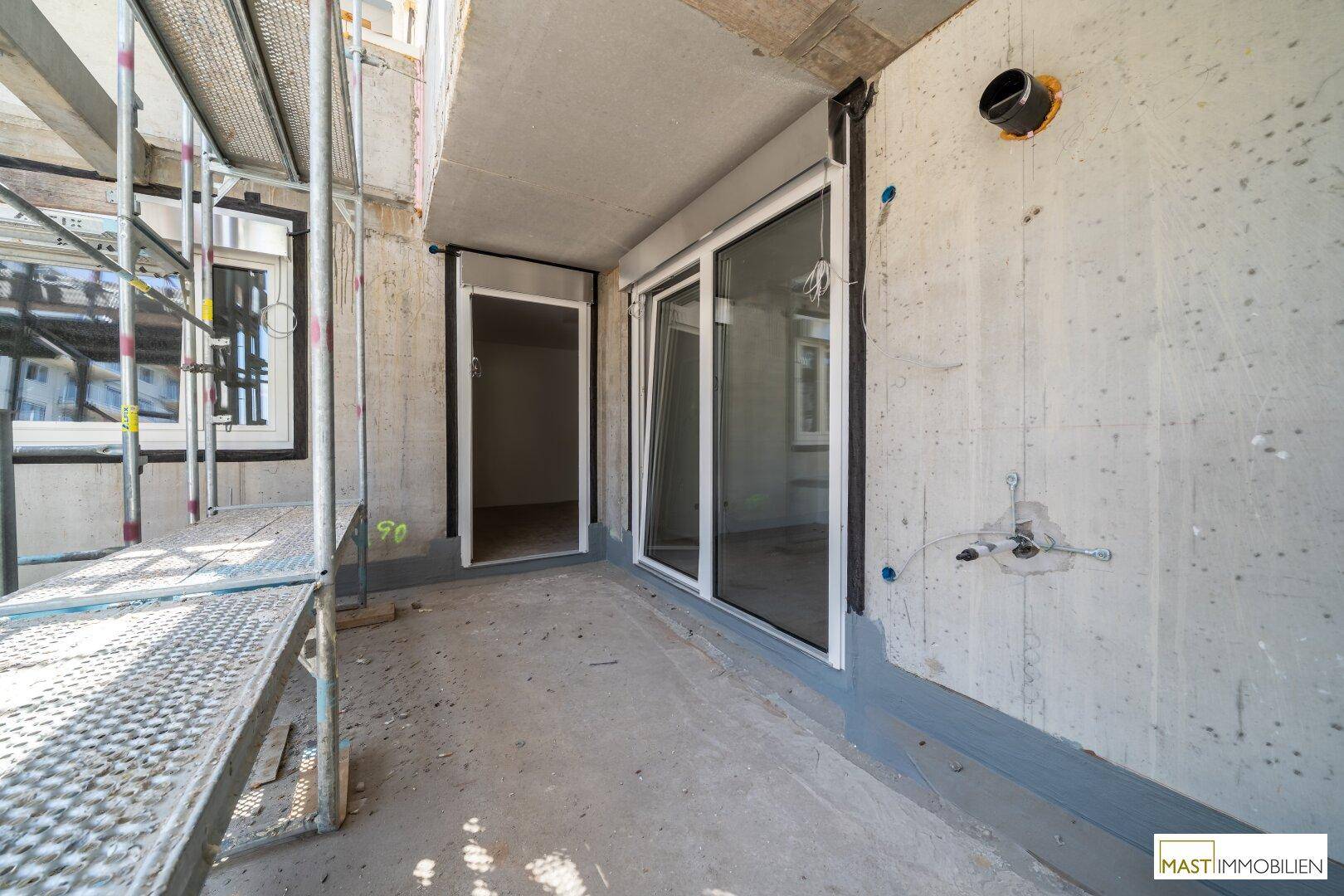 Unfertiger Balkon mit Zugangstüren zu den Innenräumen, umgeben von Baustellengerüst.