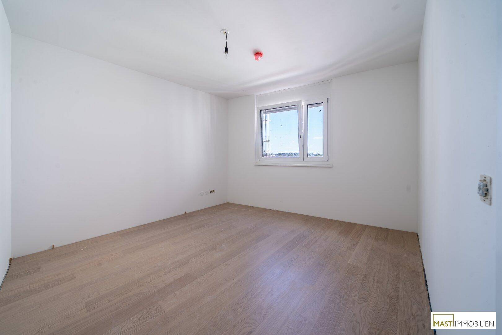 Leeres Schlafzimmer mit hellem Holzboden und einem Fenster, das viel Tageslicht hereinlässt.