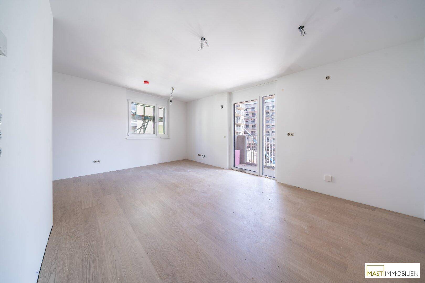 Geräumiges Wohnzimmer mit hellem Holzboden, großem Fenster und direktem Zugang zum Balkon.