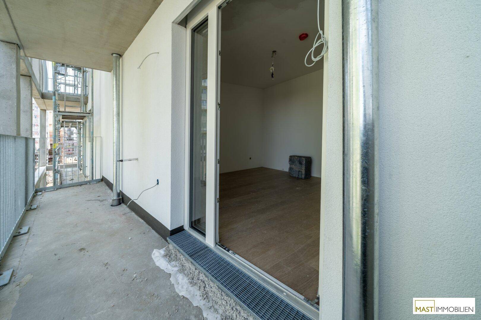Balkon mit Betonboden und Blick in einen hellen Innenraum mit Holzboden, Teil eines Neubaus.