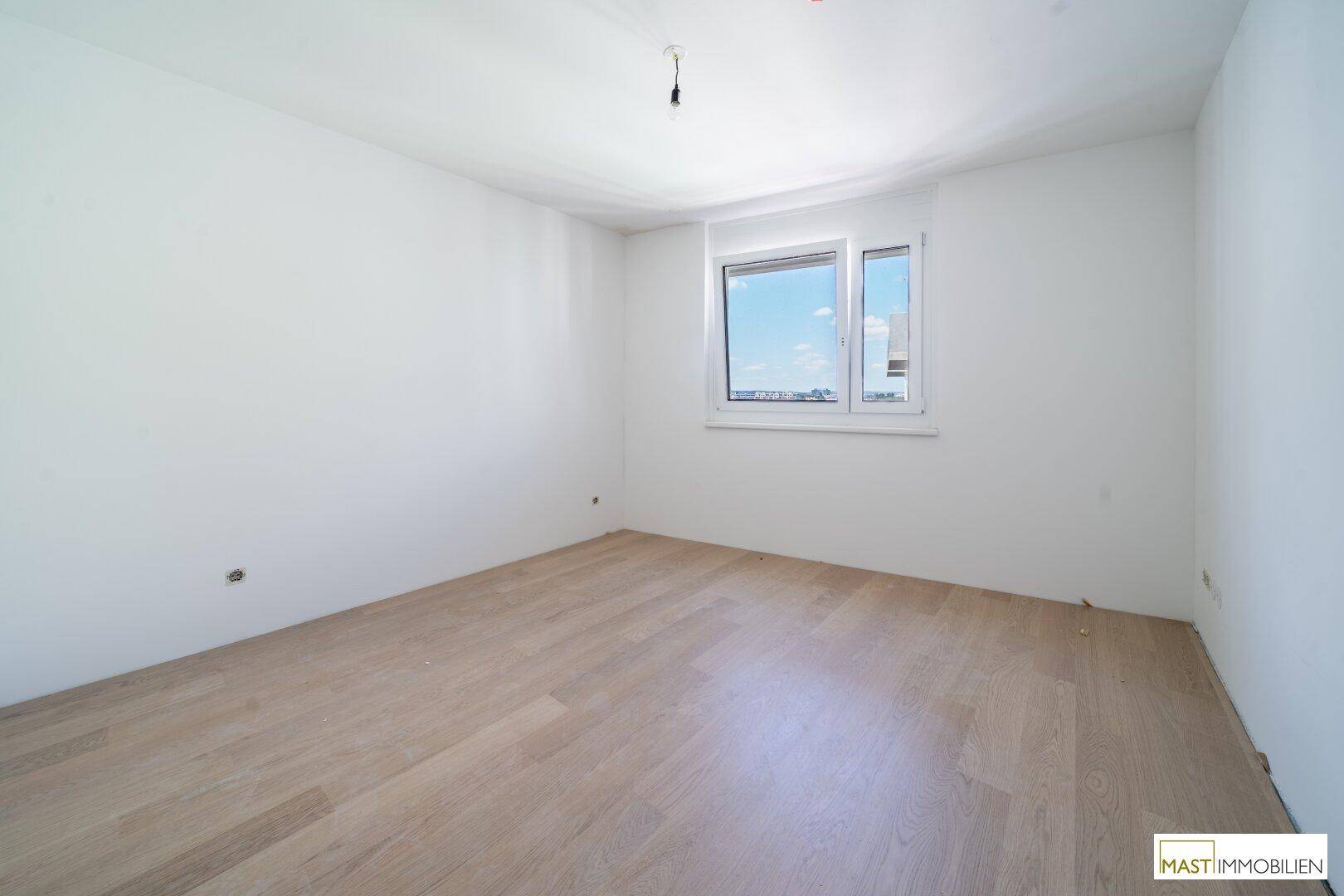 Leeres Schlafzimmer mit hellem Holzfußboden und einem großen Fenster, das viel Licht hereinlässt.