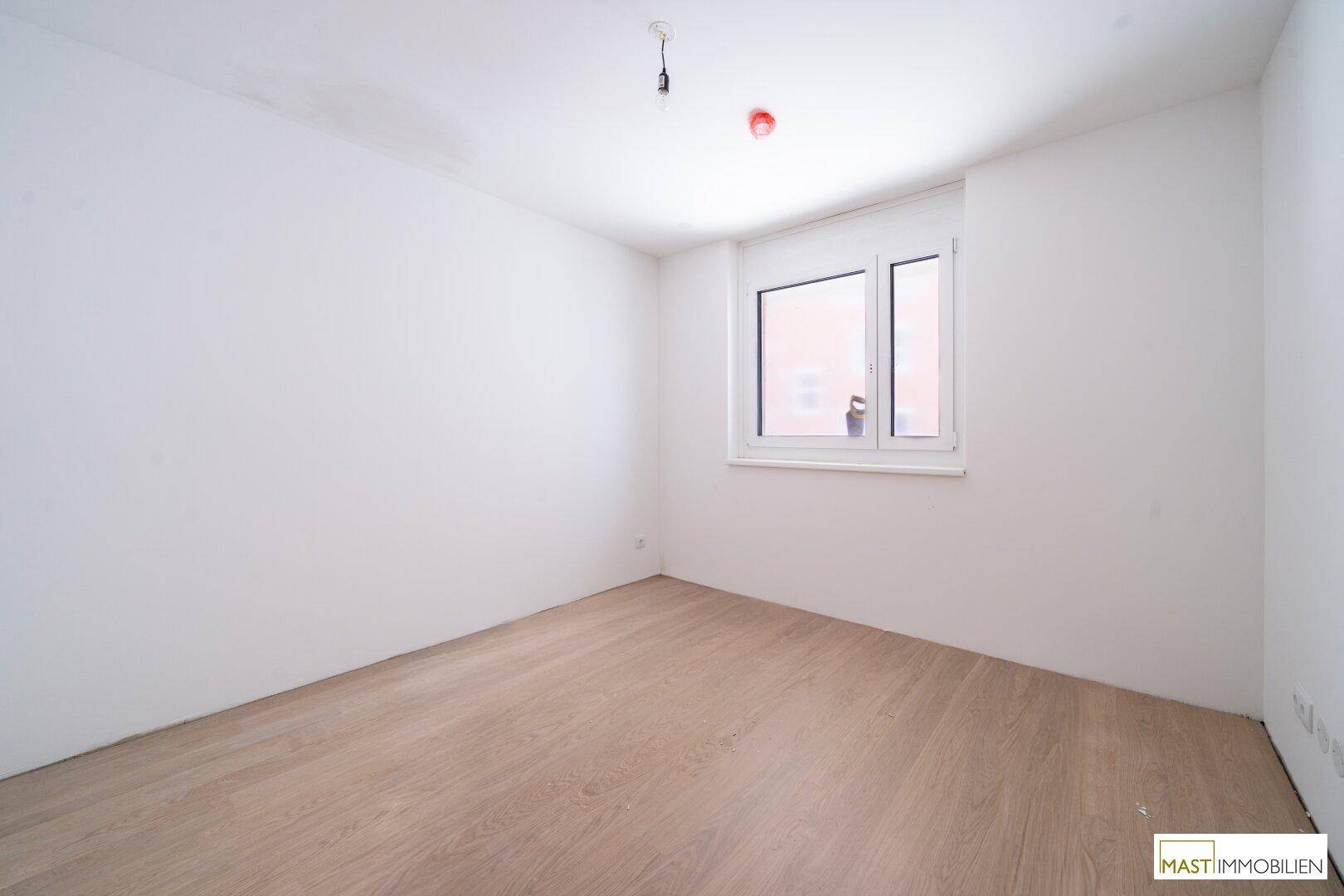 Leeres Schlafzimmer mit großem Fenster, hellem Laminatboden und frisch gestrichenen Wänden.