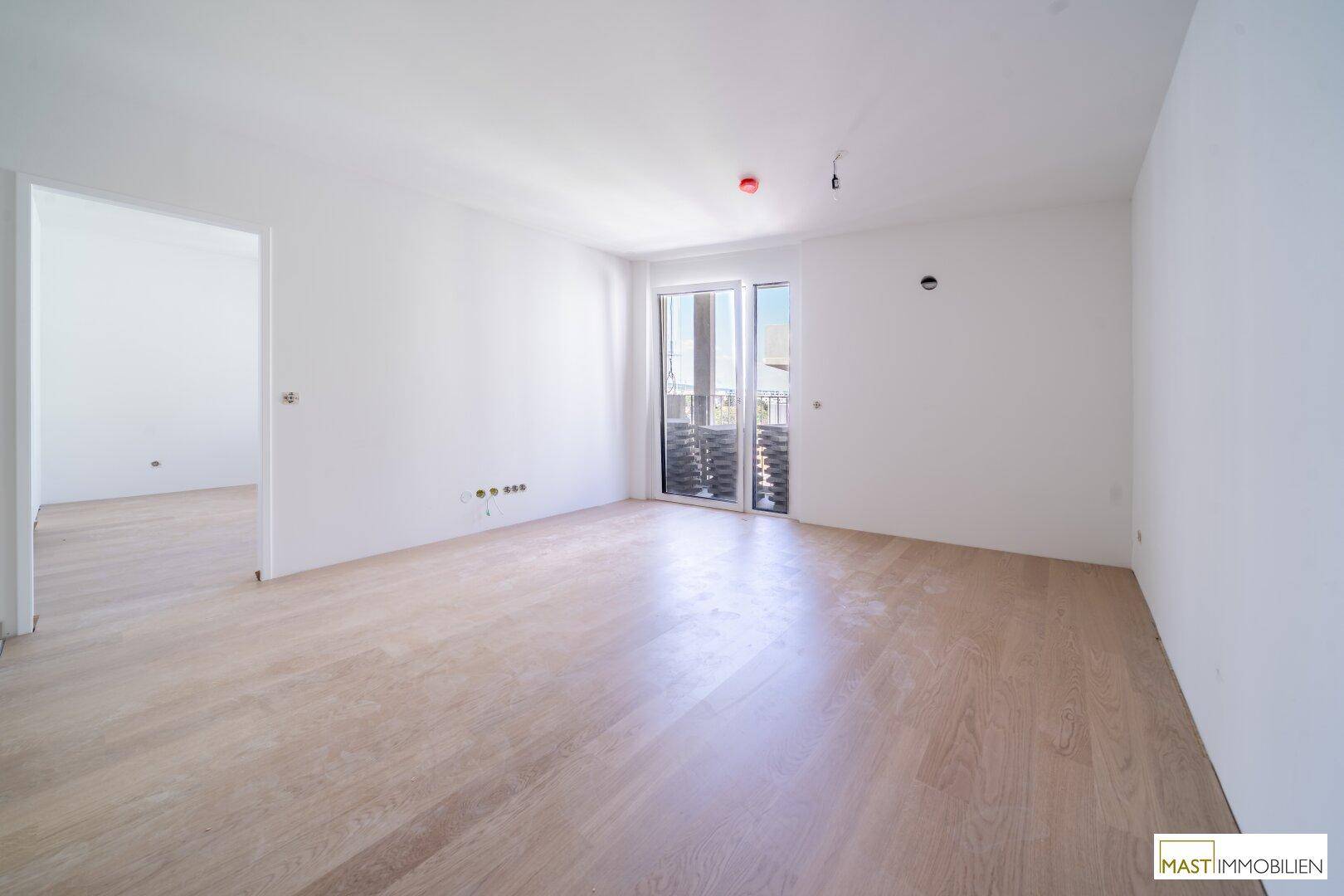 Helles, unmöbliertes Zimmer mit hellem Parkettboden und Zugang zu einem Balkon.