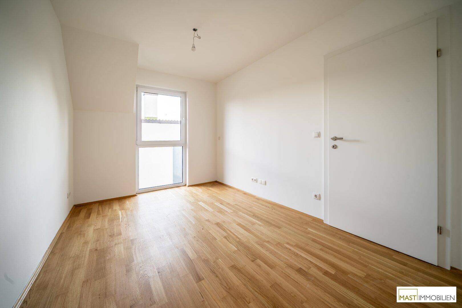 Helles Schlafzimmer mit Parkettboden und großem Fenster, das viel Tageslicht hereinlässt.