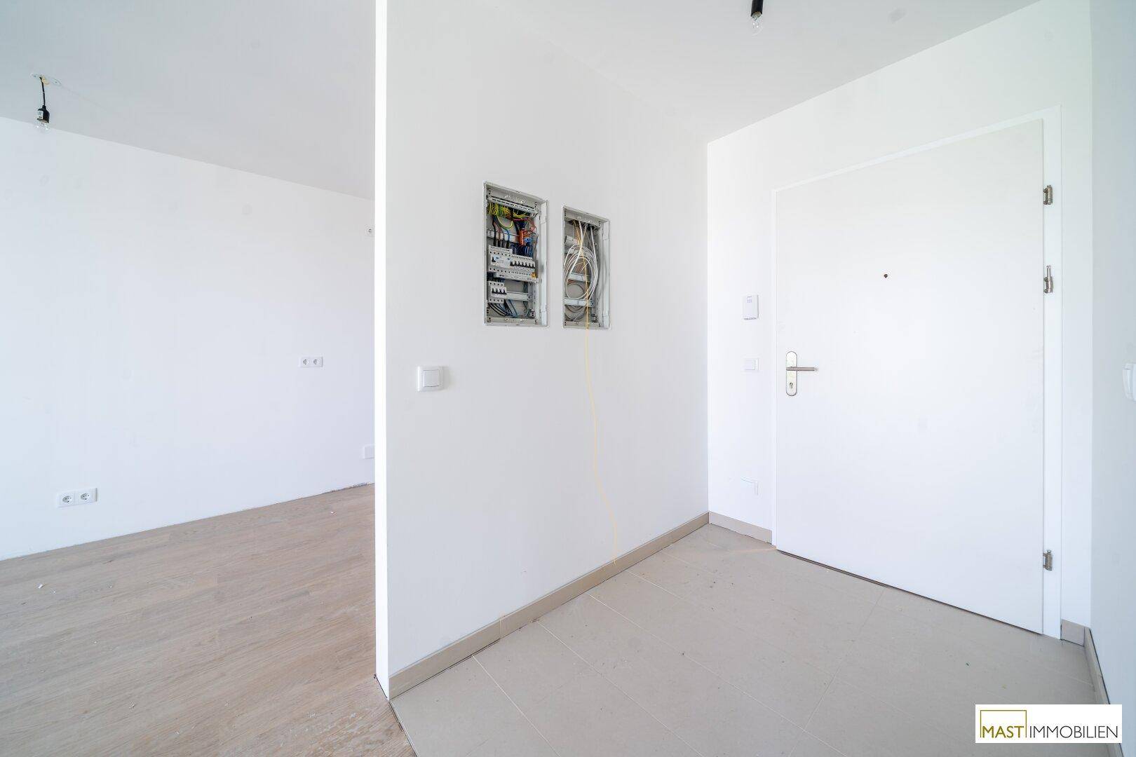 Eingangsbereich mit weißer Tür, Fliesenboden und sichtbaren Elektroinstallationen, die auf einen Neubau hinweisen.