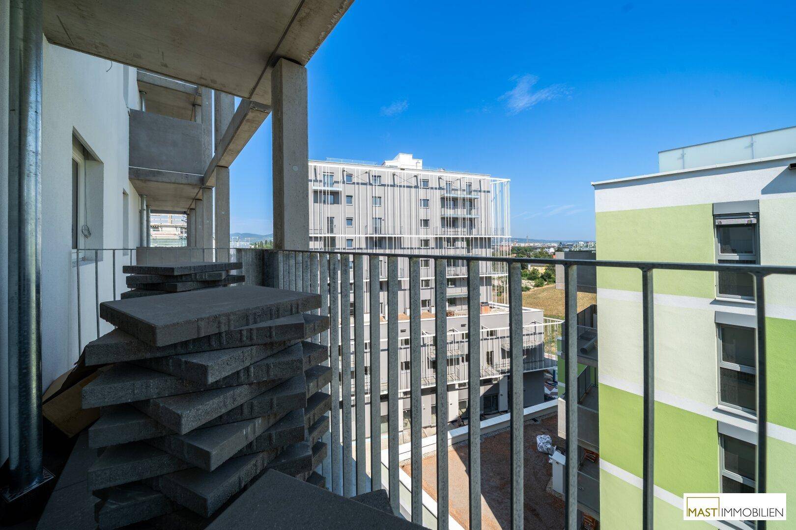 Balkon mit gestapelten Betonplatten und Blick auf umliegende moderne Wohngebäude unter blauem Himmel.