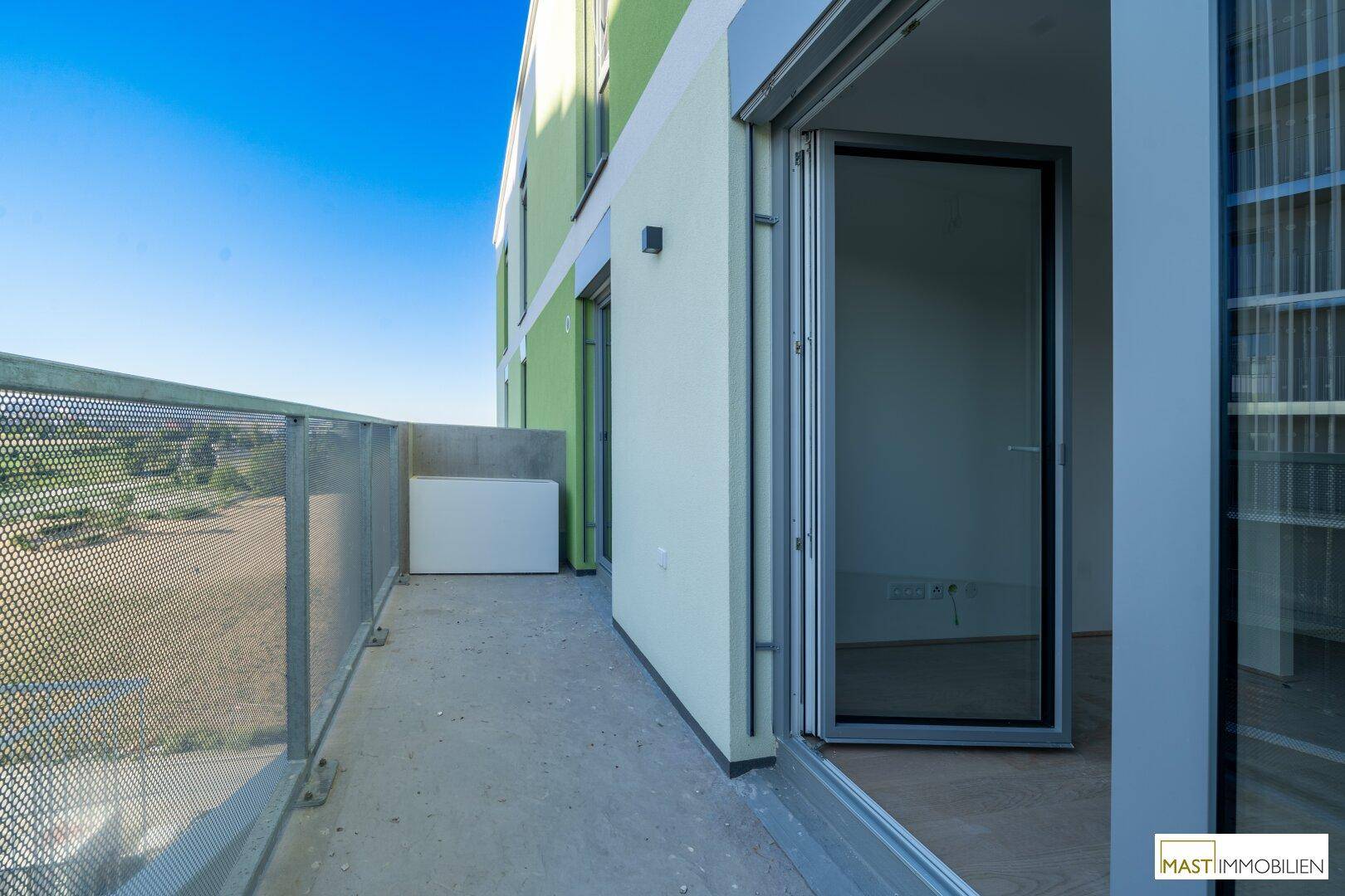 Heller Balkon mit Zugang zum Innenraum und Blick auf die grüne Umgebung und andere Gebäude.
