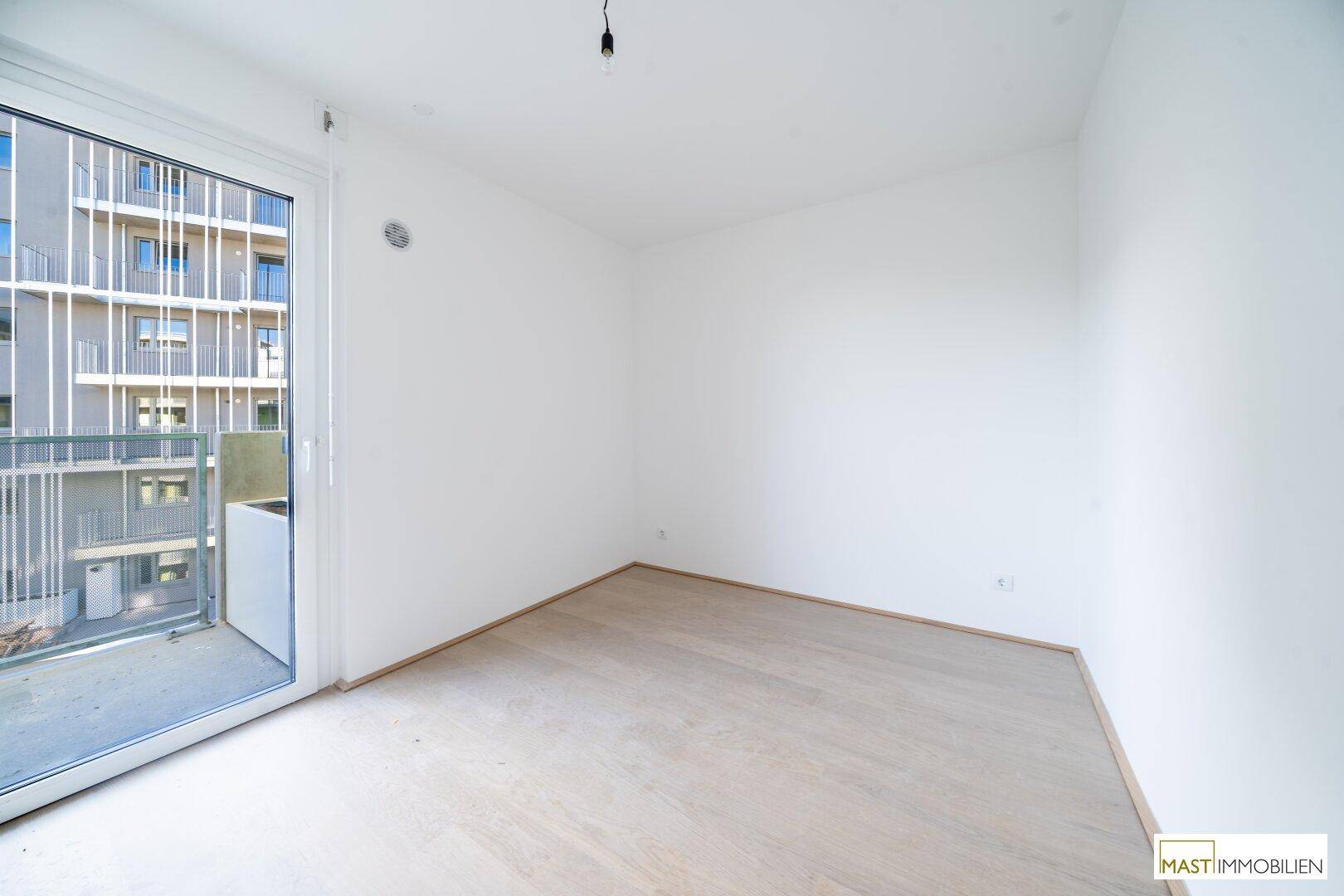 Helles Zimmer mit Parkettboden und großem Fenster, das Zugang zu einem Balkon bietet.