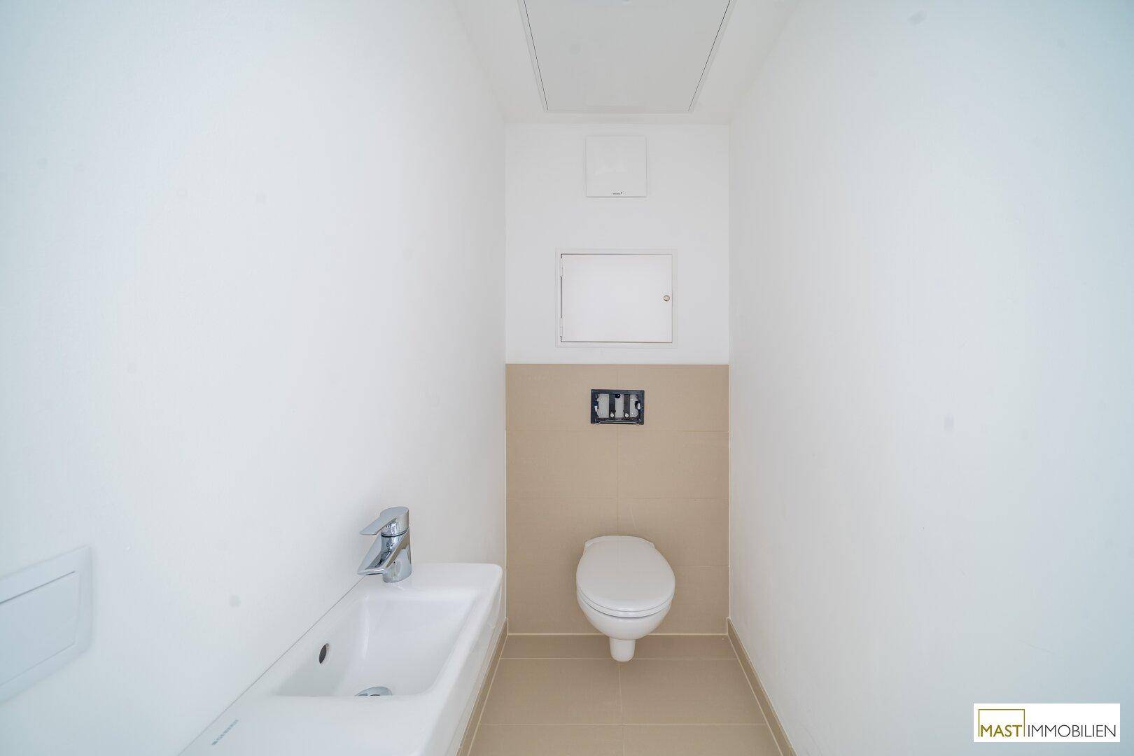Separates WC mit Wand-WC, kleinem Waschbecken und beigefarbenen Fliesen.