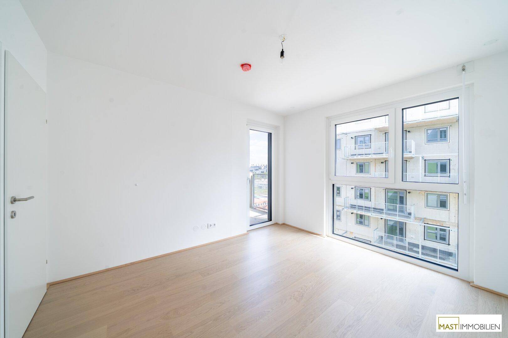 Helles Schlafzimmer mit Parkettboden und großen Fenstern mit Blick auf Nachbargebäude.