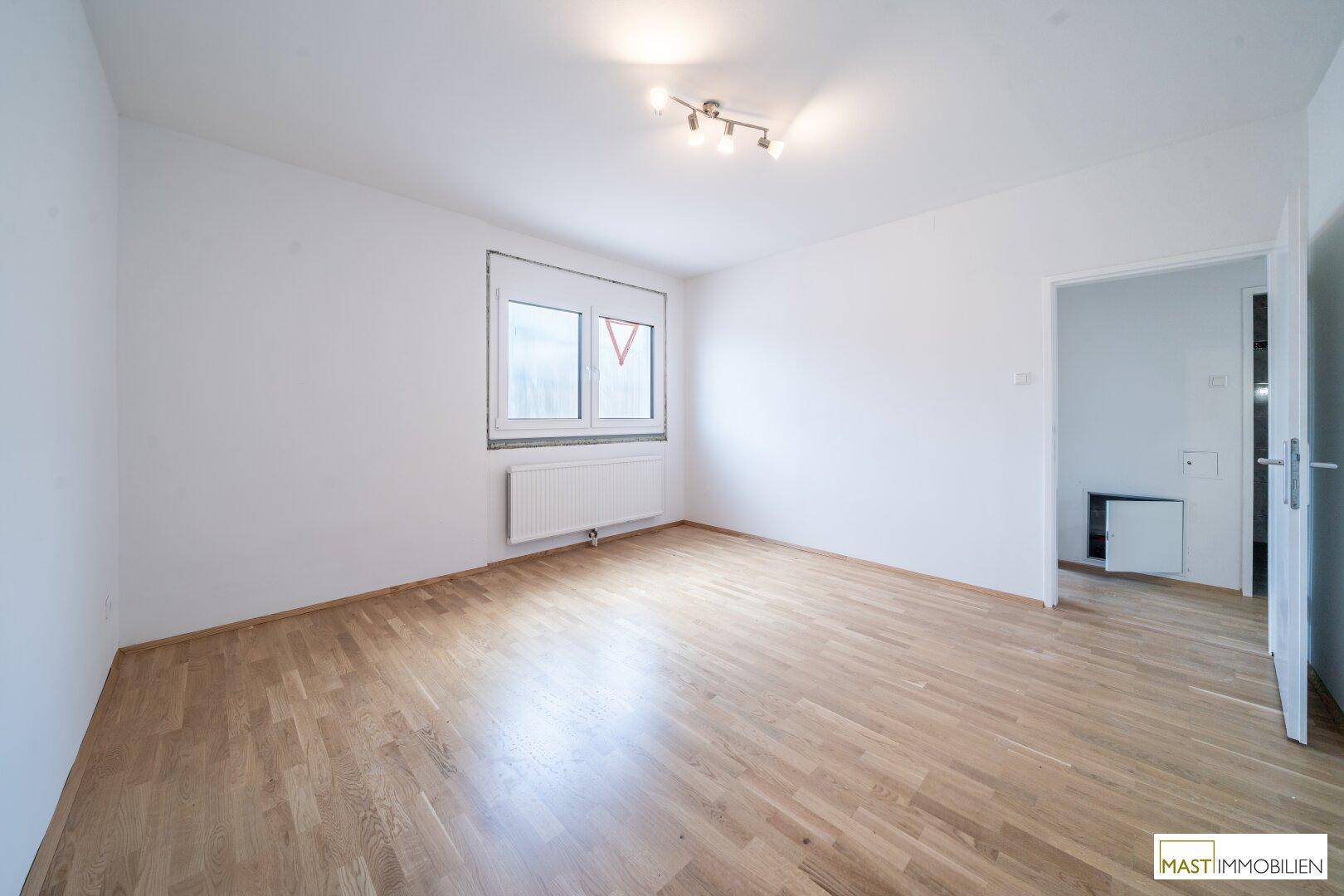 Leeres Zimmer mit Parkettboden, weißen Wänden und einem Fenster, bereit zur individuellen Gestaltung.