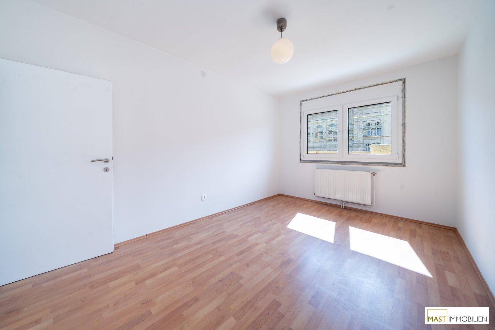 Helles Schlafzimmer mit Laminatboden und einem großen Fenster, das viel Licht hereinlässt.