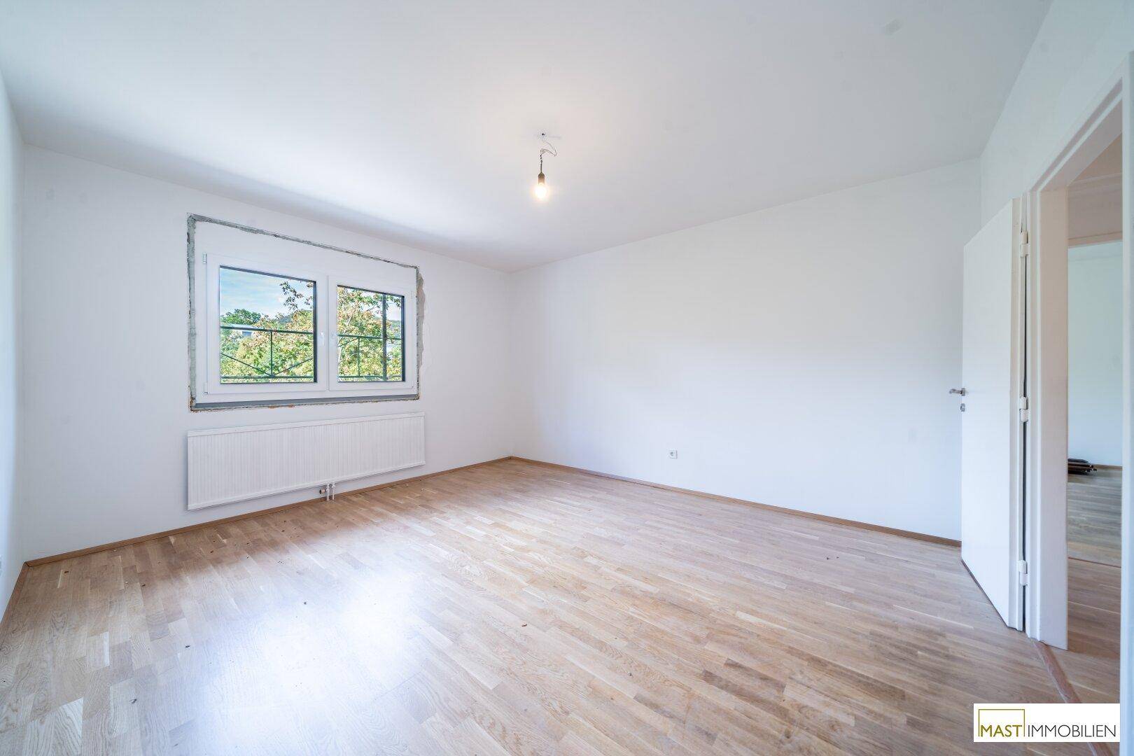 Helles, leeres Zimmer mit Holzboden und großem Fenster, ideal als Schlafzimmer.