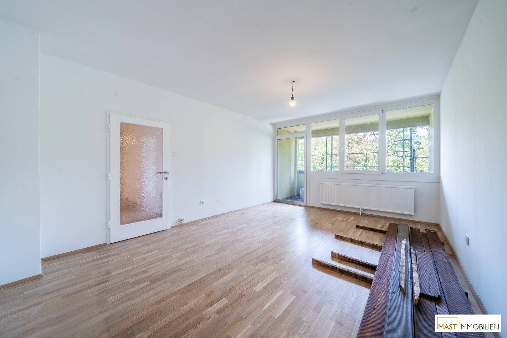 Leerer Raum mit hellem Holzboden, weißen Wänden und Zugang zu einem kleinen Balkon.