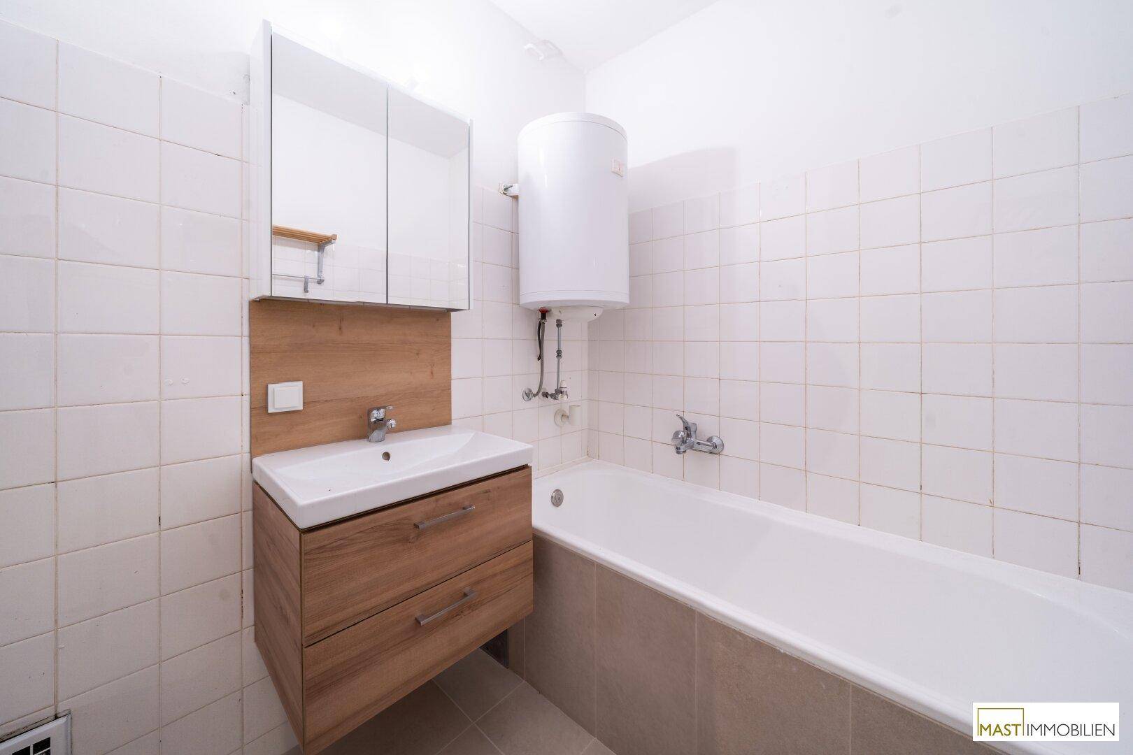 Badezimmer mit Badewanne, Waschbeckenunterschrank und weißen Fliesen an den Wänden.