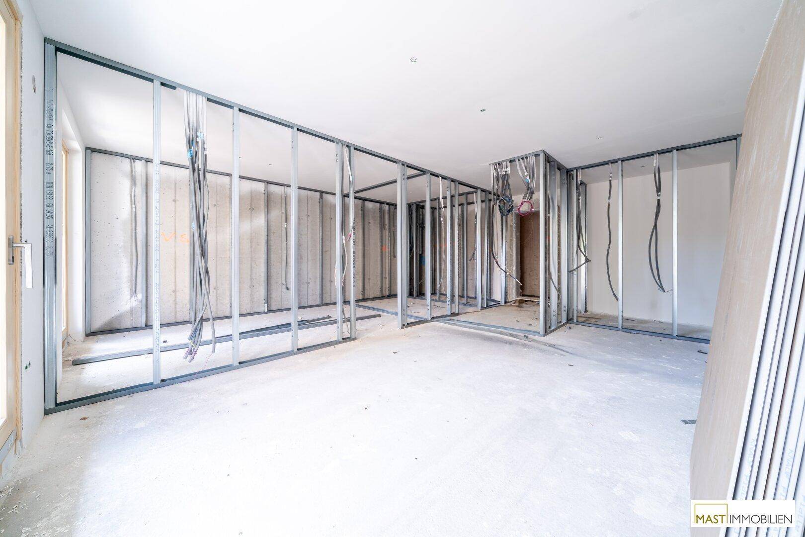 Innenraum im Rohbau mit Metallständerwerk, sichtbaren Leitungen und Betonboden.
