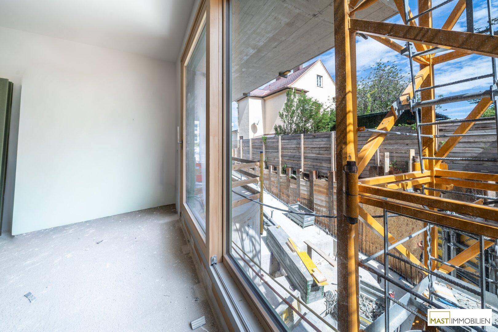 Blick aus einem unfertigen Raum durch große Fenster auf die Baustelle mit Gerüst und einem angrenzenden Gebäude.
