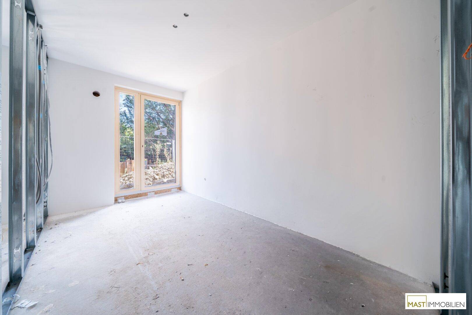 Leerer Raum im Rohbauzustand mit Betonboden und einer Glastür, die ins Freie führt.