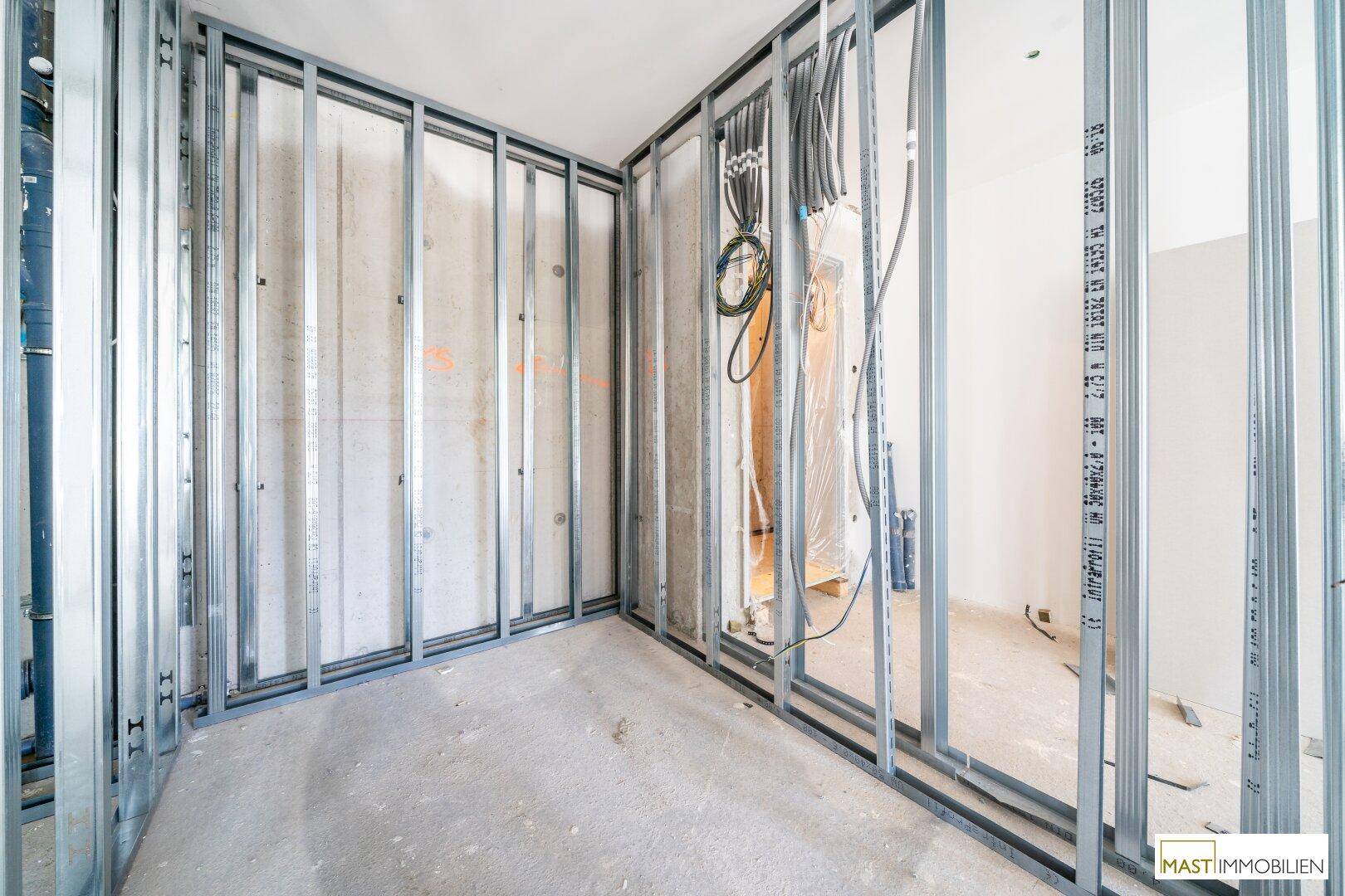 Ecke eines Rohbauraumes mit Metallständerwerk, sichtbaren Leitungen und Betonwand.