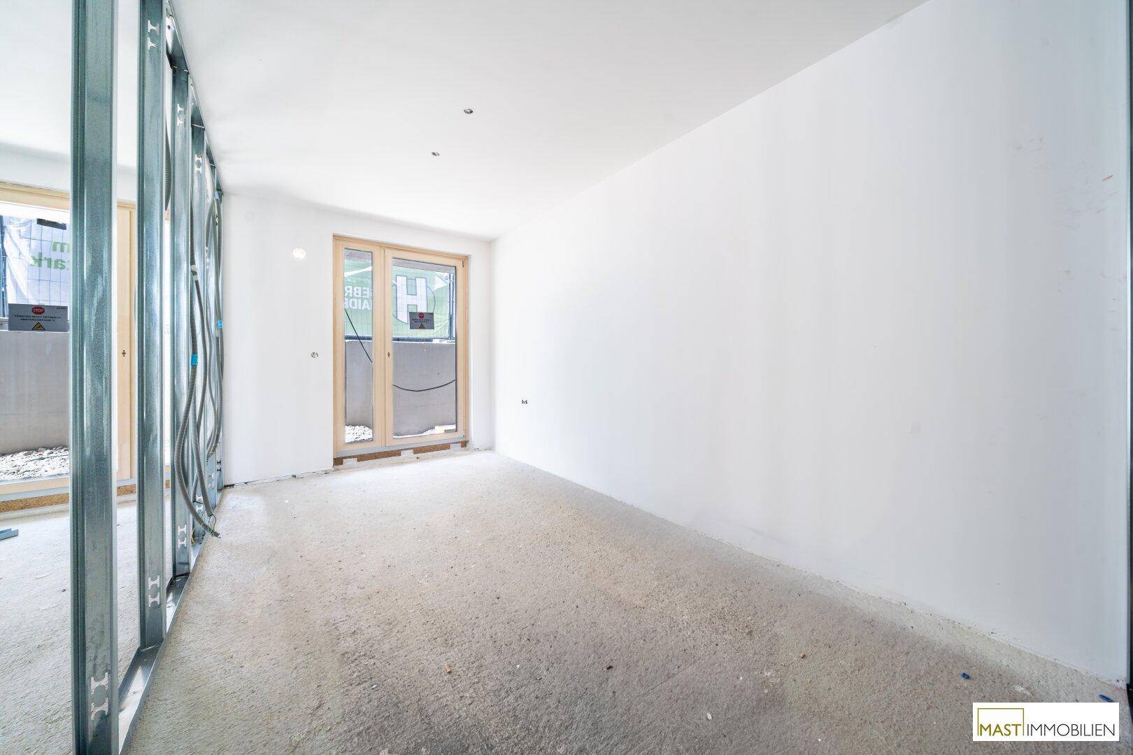 Unfertiger Raum mit Betonboden, weißen Wänden und Zugang zu einem Außenbereich während der Bauphase.