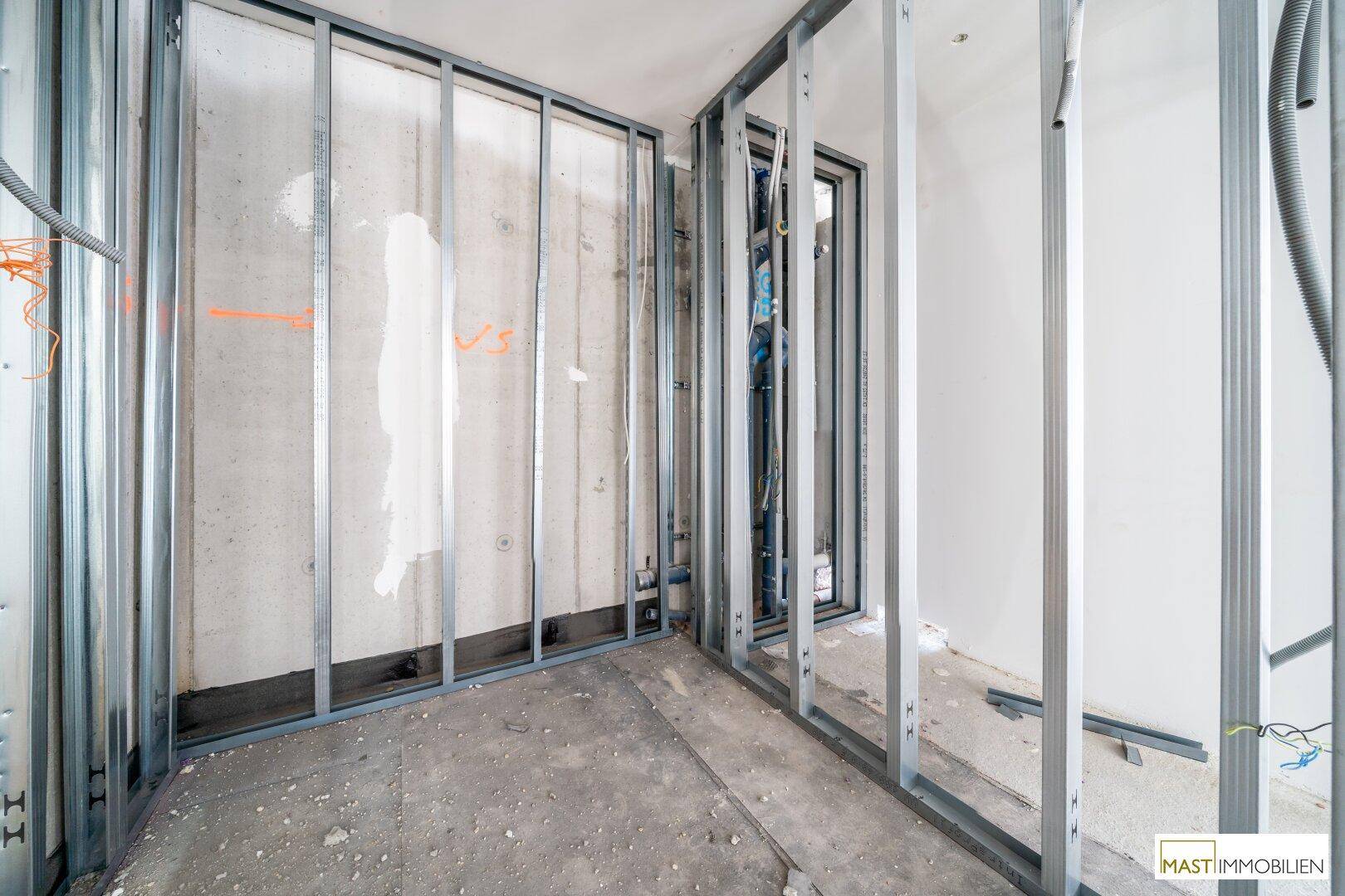 Unfertiger Innenraum mit Betonboden, weißen Wänden und Metallständerwerk, das eine Ecke bildet.