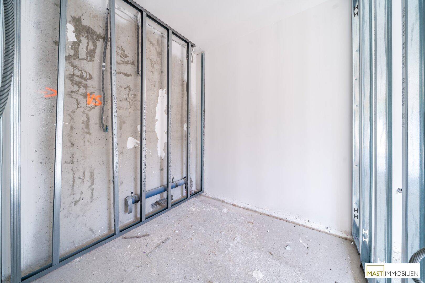 Rohbau eines Raumes mit Betonboden, weißen Wänden und sichtbaren Installationen für Sanitäranlagen.