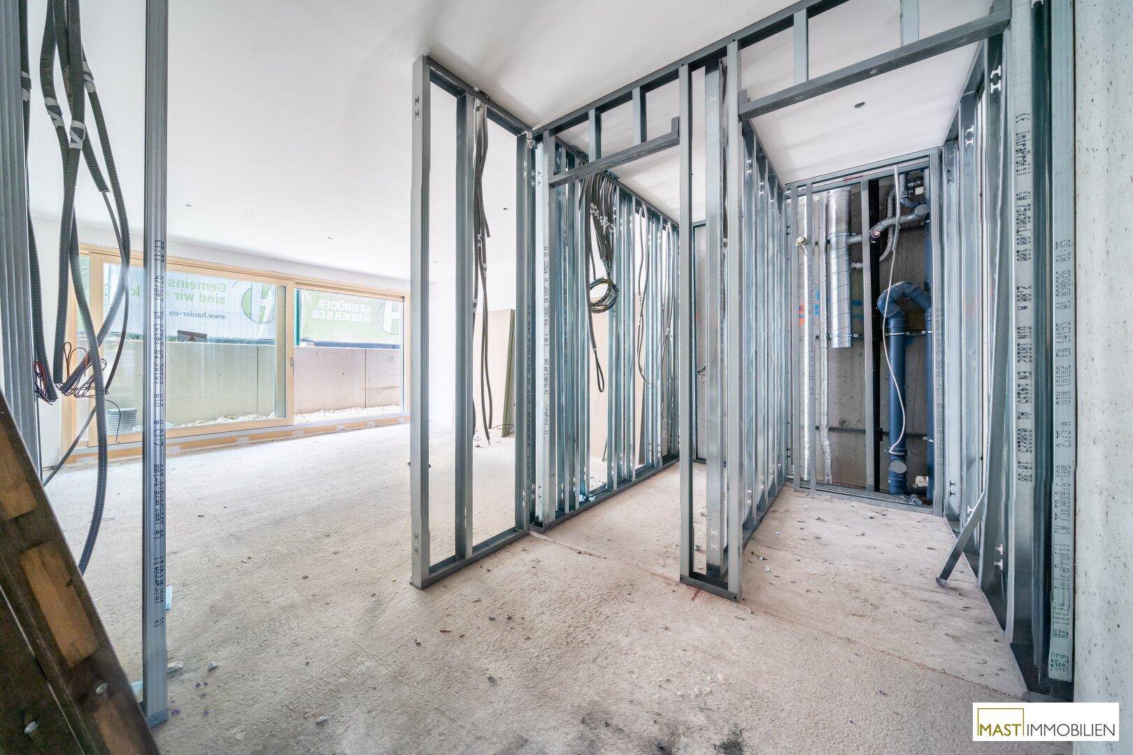 Rohbau-Bereich mit Metallständerwänden, sichtbaren Leitungen und großen Fenstern im Hintergrund.
