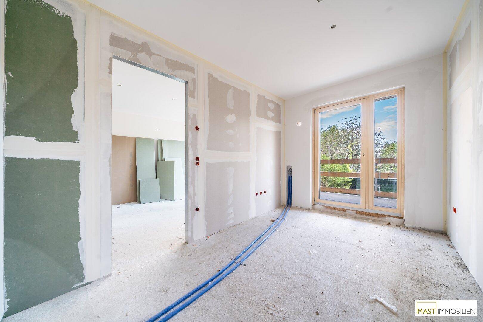 Unfertiger Raum mit Trockenbauwänden, großen Fenstern und sichtbaren Installationen, bereit für den Innenausbau.