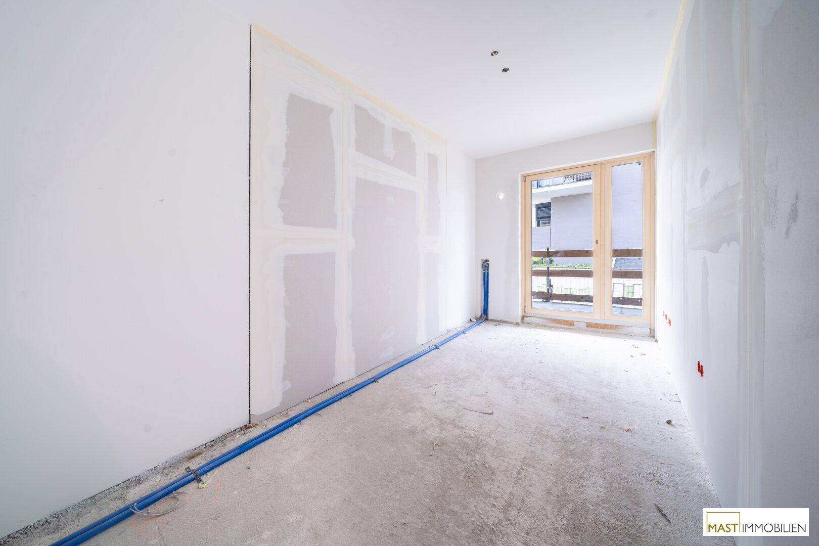 Rohbauzimmer mit sichtbaren Installationen und Fenster zur Außenseite.