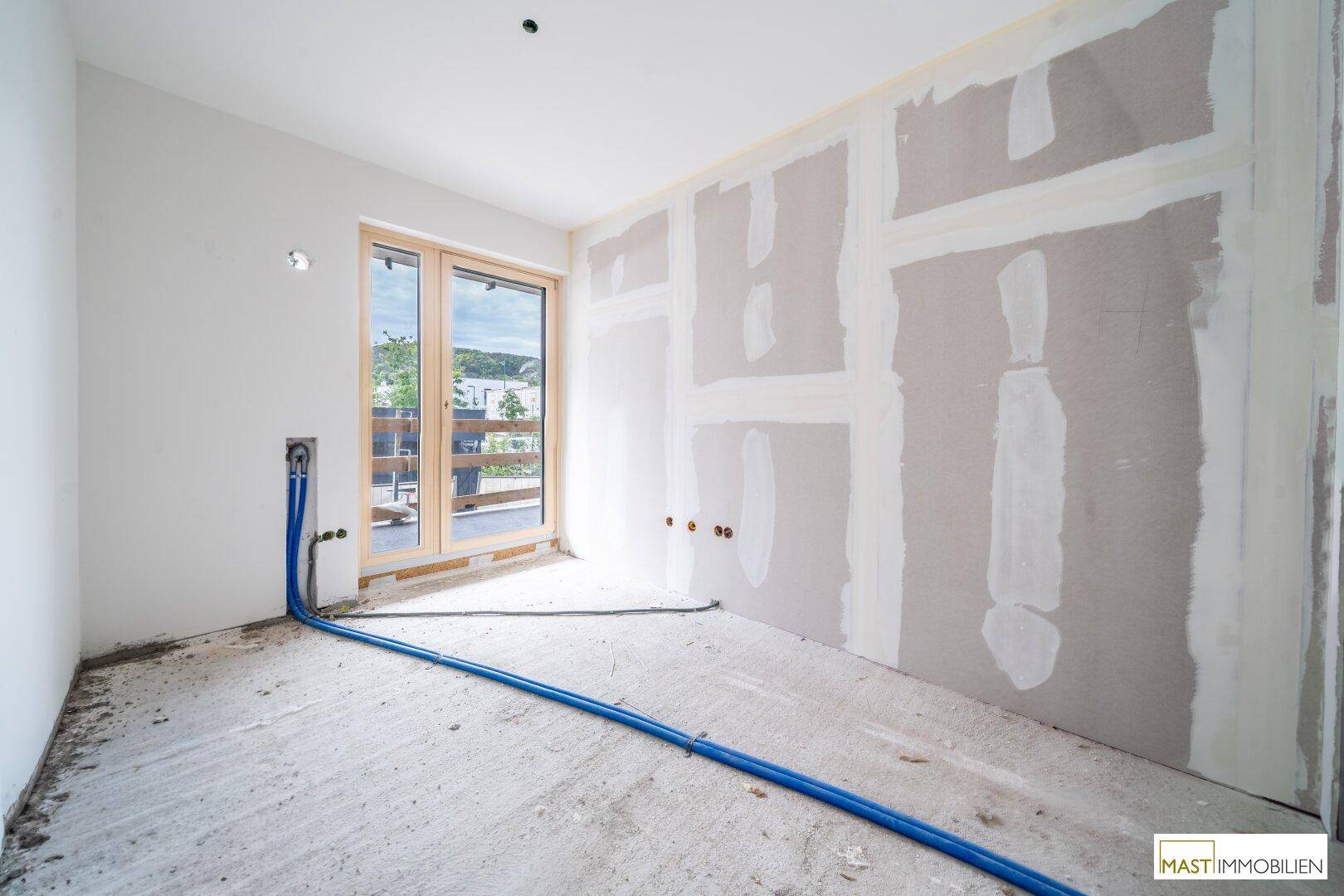 Rohbauzimmer mit vorbereiteten Sanitäranschlüssen und Zugang zu einem Außenbereich durch eine Holztür.