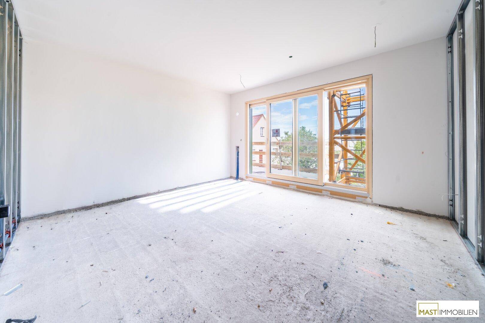 Unfertiger Innenraum mit großen Fenstern und Blick auf die Baustelle im Freien.