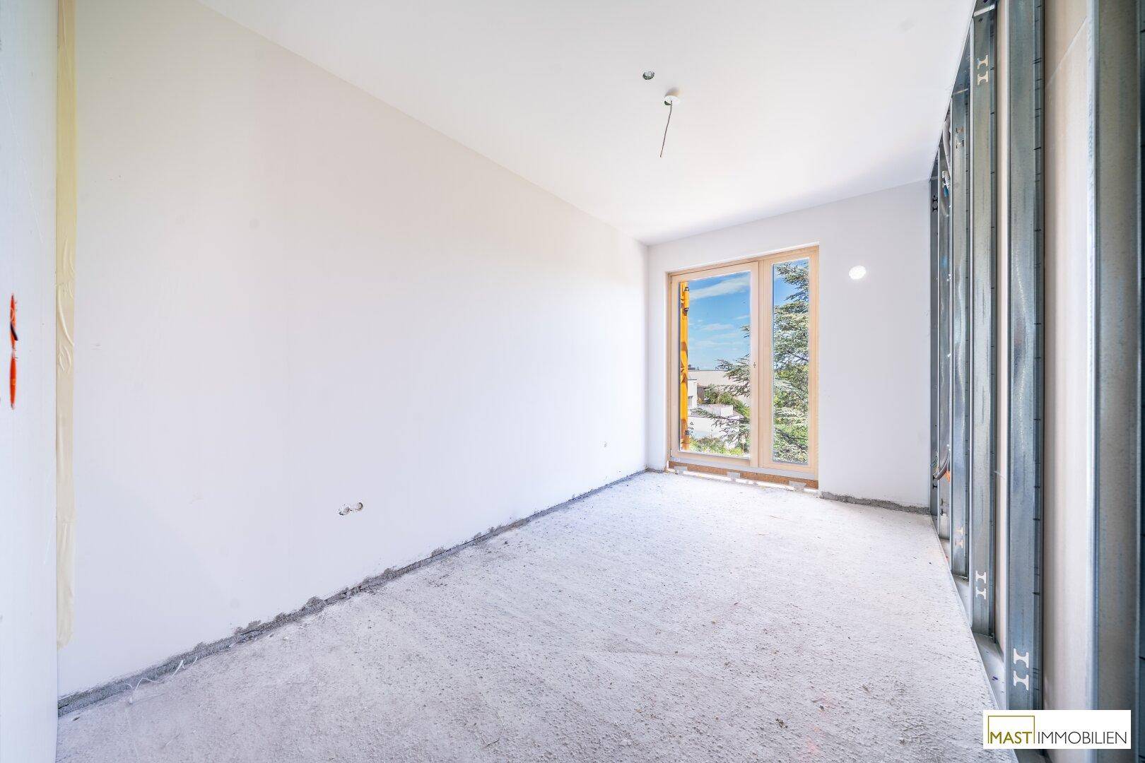 Unfertiges Schlafzimmer mit weiß gestrichenen Wänden, Betonboden und einem Fenster mit Blick ins Grüne.