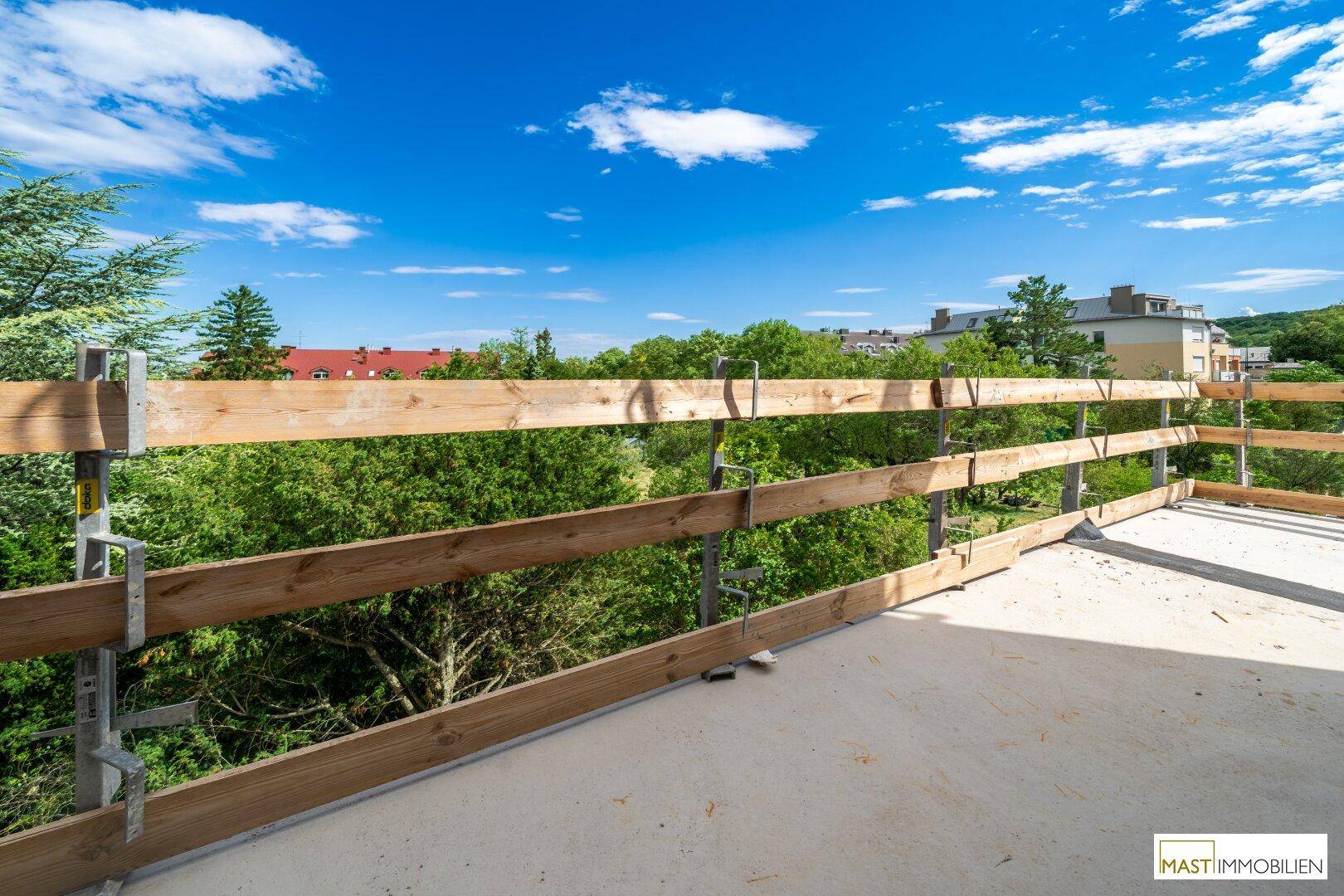 Unfertiger Balkon mit temporärem Holzgeländer und weitem Blick auf grüne Bäume und blauen Himmel.