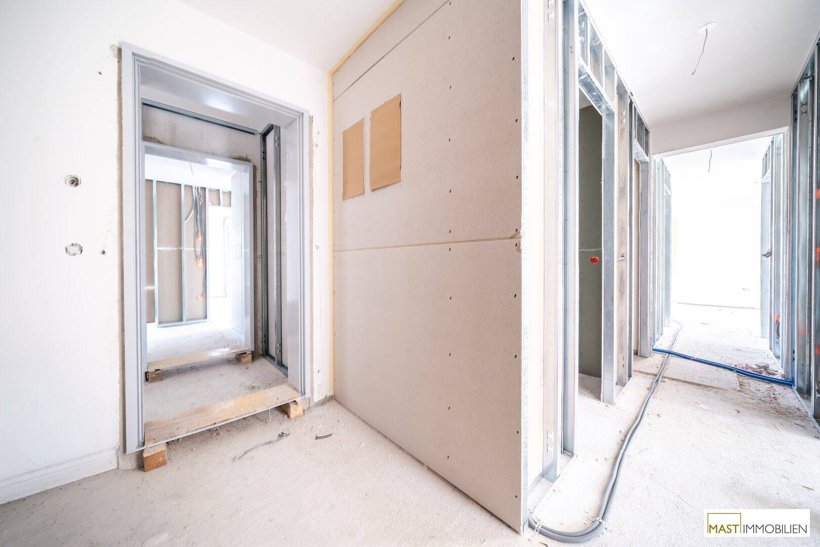 Rohbau-Flur mit Türrahmen und Metallständern, der den Übergang zu weiteren Räumen zeigt.