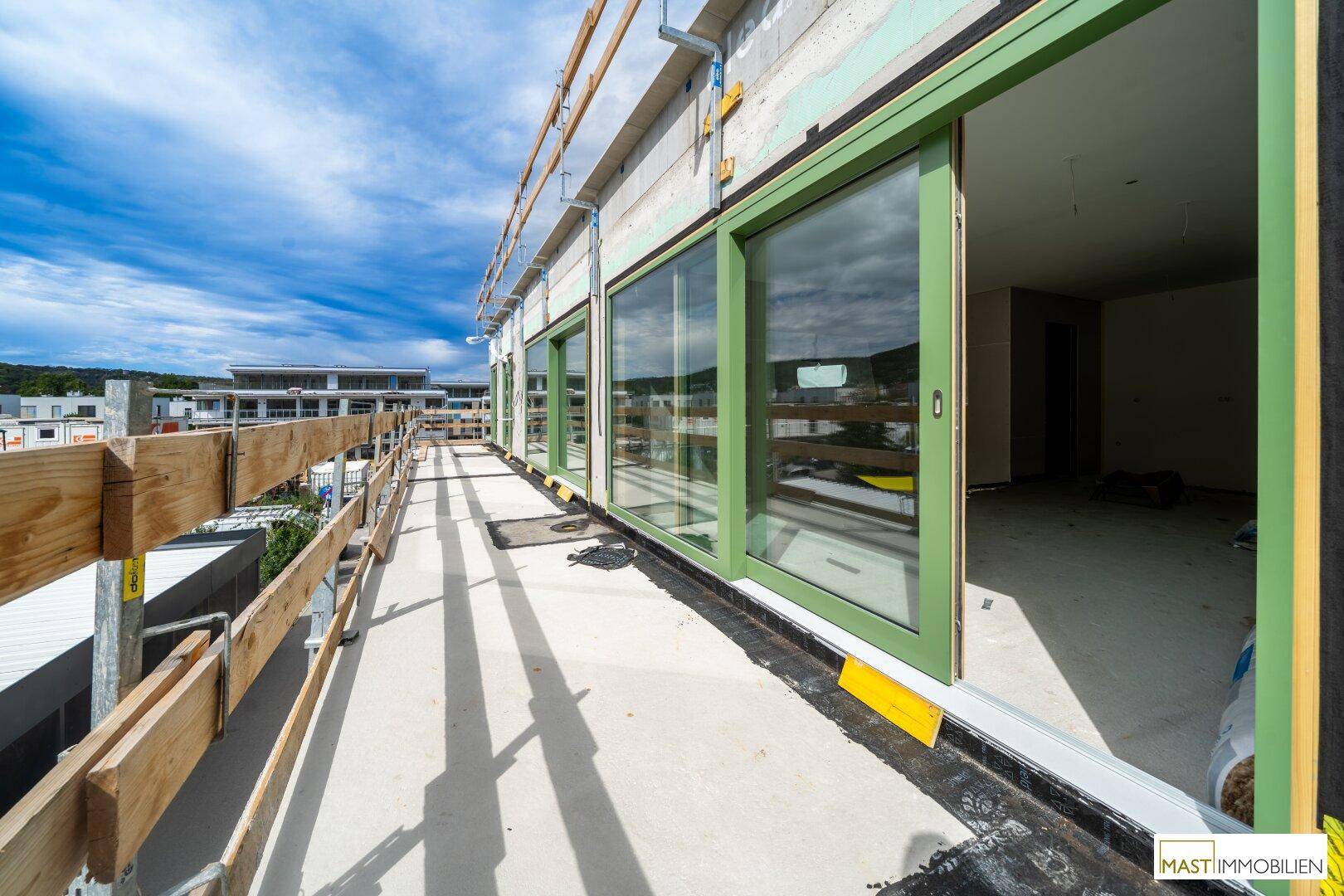 Langer Balkon im Rohbau mit Holzgeländer und großen grünen Schiebetüren.