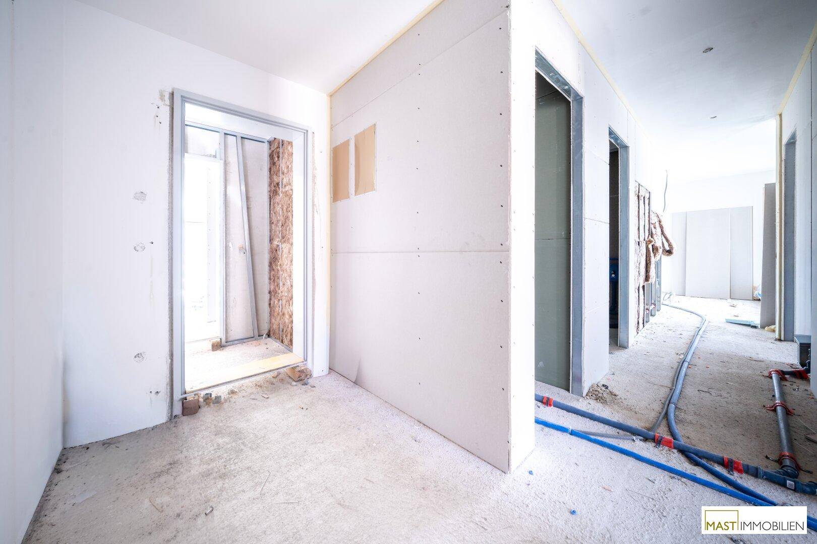 Flur im Rohbauzustand mit Trockenbauwänden und sichtbaren Installationen, der zu weiteren Räumen führt.