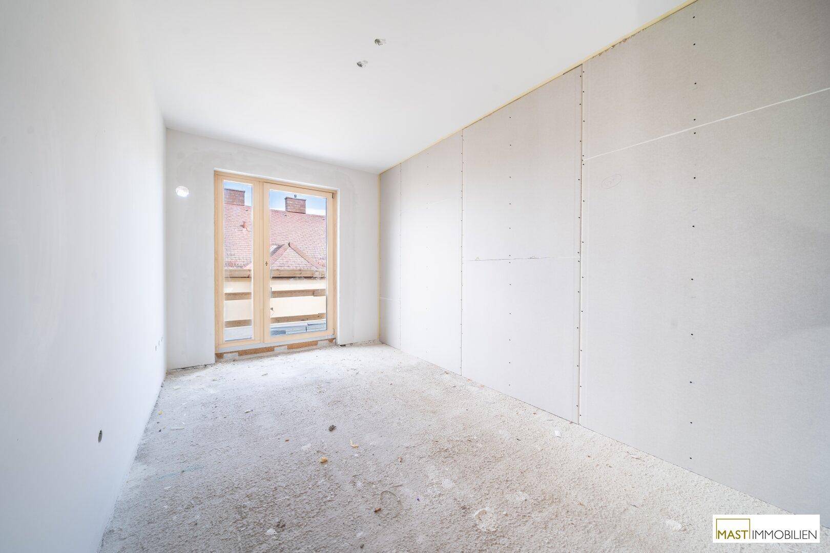 Rohbau-Zimmer mit Trockenbauwänden und einem Fenster, das auf ein Dach blickt, bereit für den Ausbau.