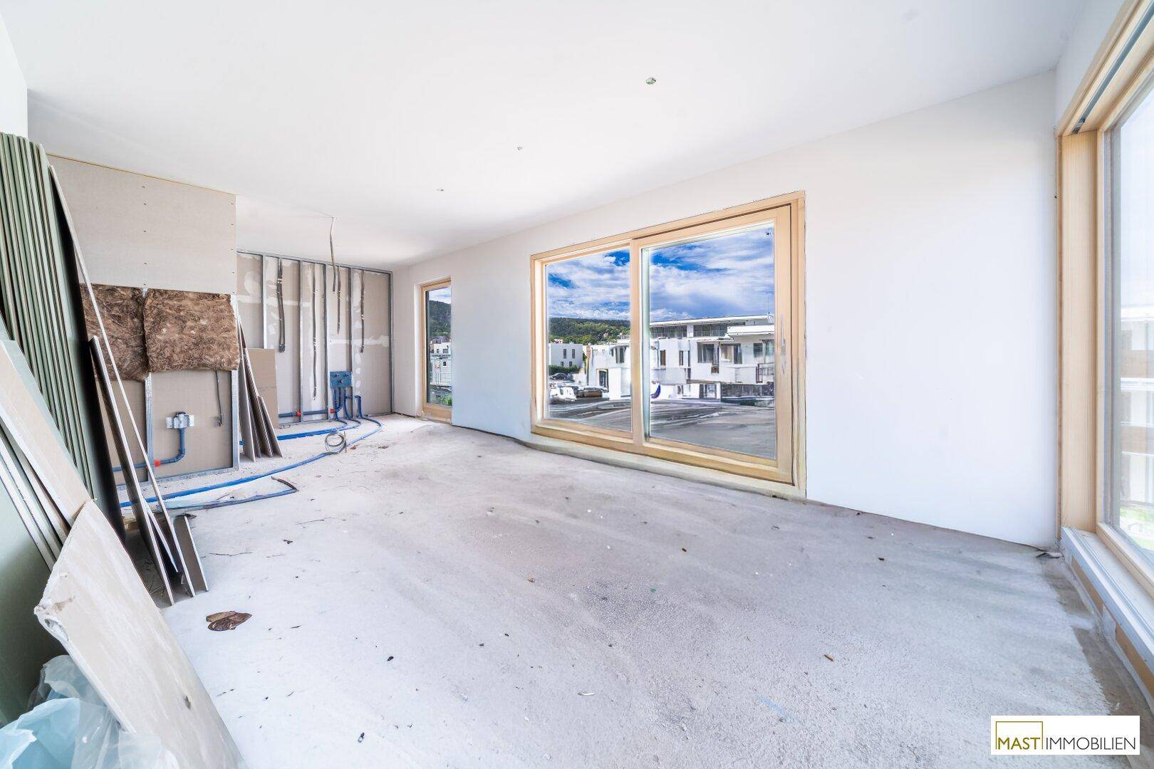 Geräumiger Rohbau-Raum mit großen Fenstern, Blick nach draußen und sichtbaren Installationsvorbereitungen.