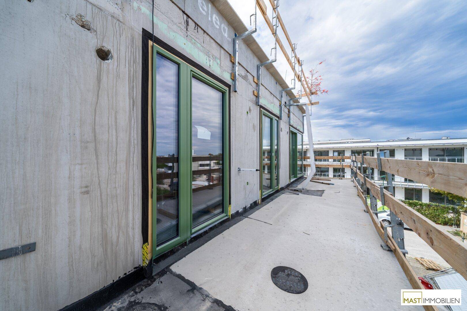 Im Bau befindlicher Balkon mit grünen Fensterrahmen und Blick auf die Baustelle und umliegende Gebäude.